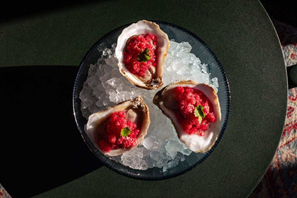Evangeline Rooftop | Overhead shot of three oysters in a bowl sitting on ice dressed with pink toppings and a parsley leaf | Homes Almanac