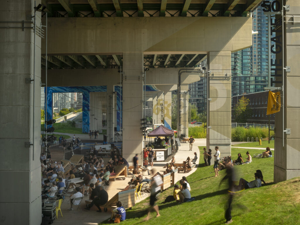 Beneath the Gardiner at The Bentway | A sunny day with people walking around, and sitting on a grassy hill | Homes Almanac