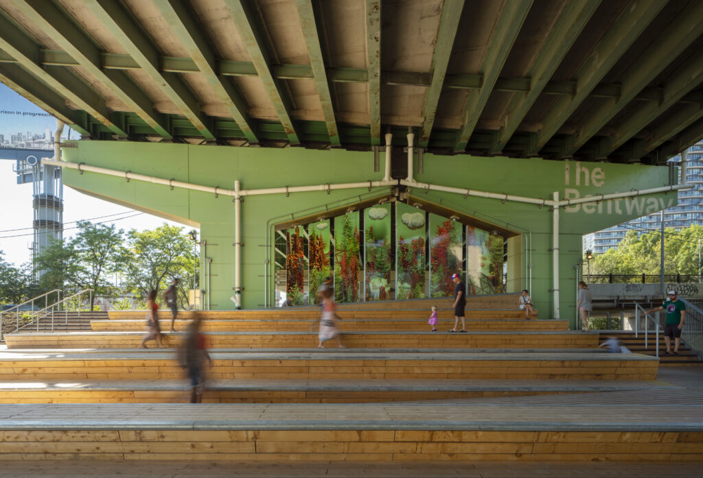Beneath the Gardiner at The Bentway | Soft daylight coming in with green painted concrete columns and people walking along wooden steps | Homes Almanac