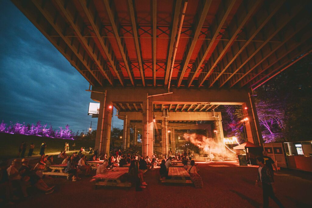 Beneath the Gardiner at The Bentway | Nighttime with people sitting at picnic tables watching a show turning the area a bright red while purple lighting lights up trees | Homes Almanac