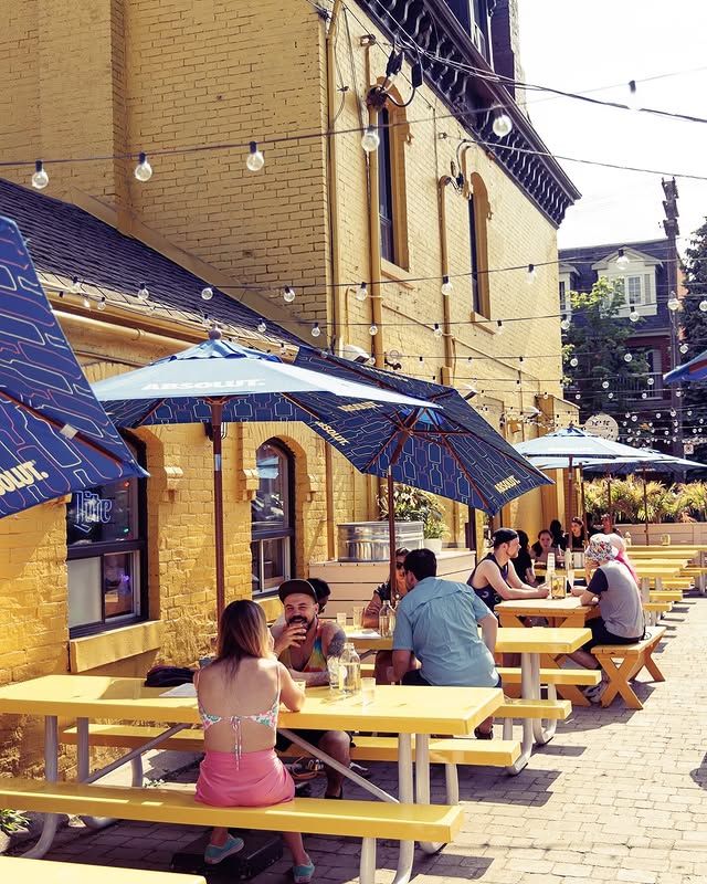 Pennies Toronto | Bright yellow brick building with dark blue patio umbrellas and people sitting on yellow picnic tables | Homes Almanac