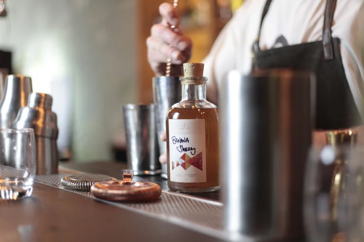 fermentation cocktails toronto | A bottle of fermented liquid with a hand stirring a cocktail in the background | Homes Almanac