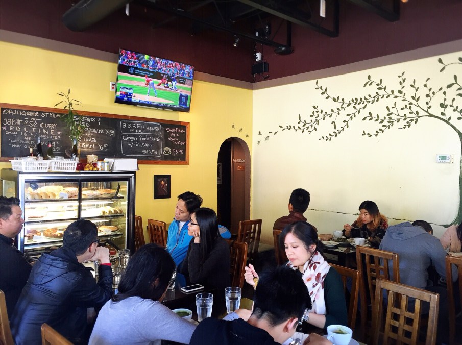 Cafe de l'Orangerie French Japanese Cafe Vancouver | Interior photo of restaurant guests sitting at tables on wooden chairs, a TV screen showing baseball, and menu features written on a chalkboard | Homes Almanac