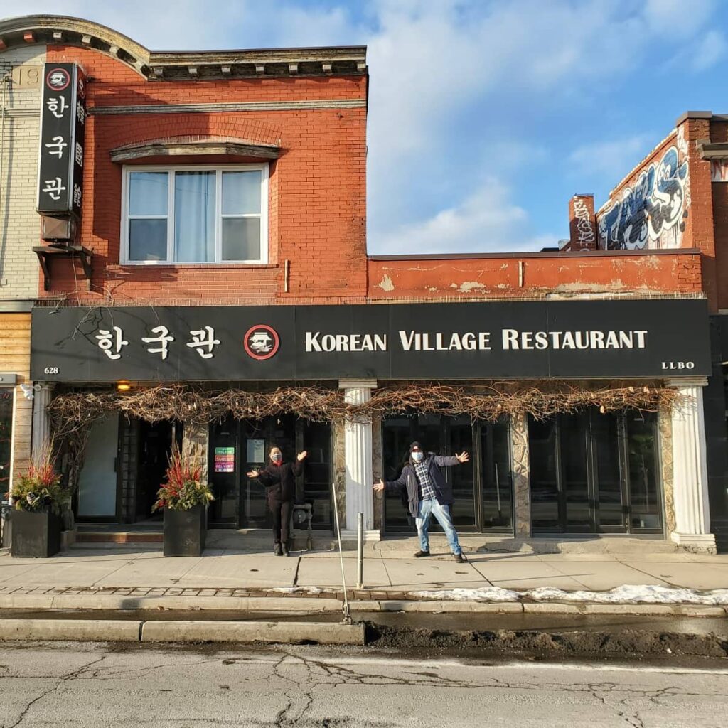 Exterior shot of Korean Village Restaurant with a black sign and Jason and his wife standing outside on a sunny day | Korean Village in Koreatown | Homes Almanac