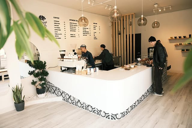 Ninetails Coffee Bar | A sleek white coffee bar with two baristas in black behind the counter and a customer ordering | Homes Almanac