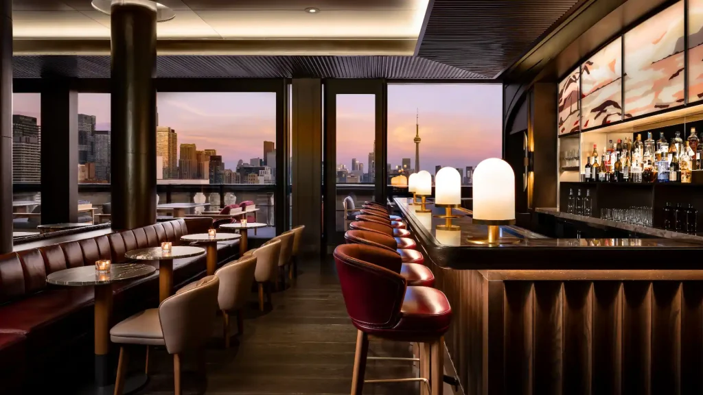 Park Hyatt Toronto | Sunset shot of a rooftop bar with floor to ceiling windows, a bar to the right with lush red bar stools and table seating to the left | Homes Almanac