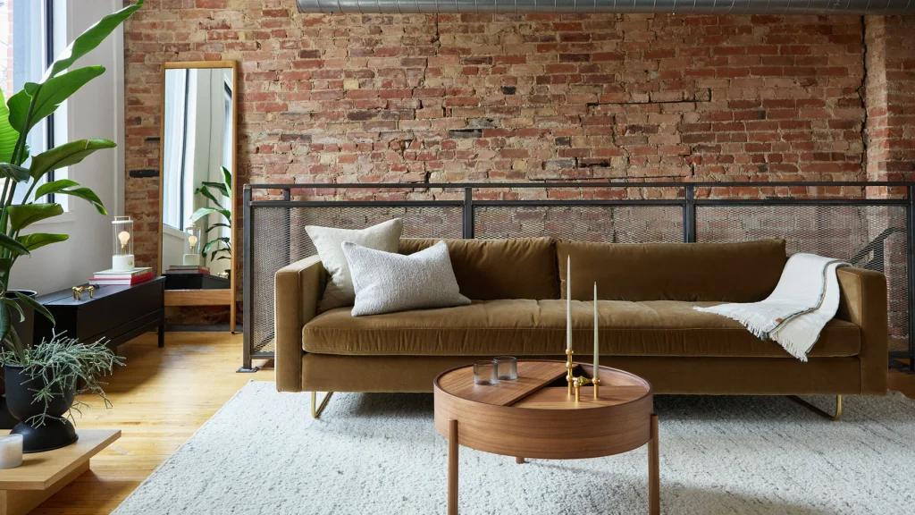 Ossington Design & Home Goods | Daylight filled room with an olive green suede couch on a grey carpet with brown circle coffee table and a brick wall in the background | Homes Almanac
