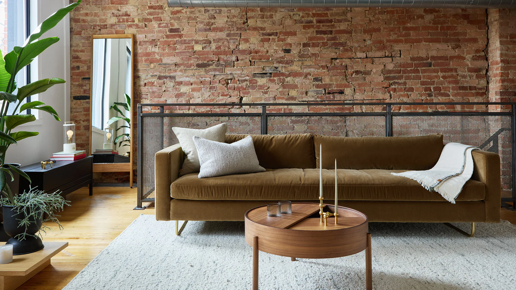 Ossington Design & Home Goods | Daylight filled room with an olive green suede couch on a grey carpet with brown circle coffee table and a brick wall in the background | Homes Almanac
