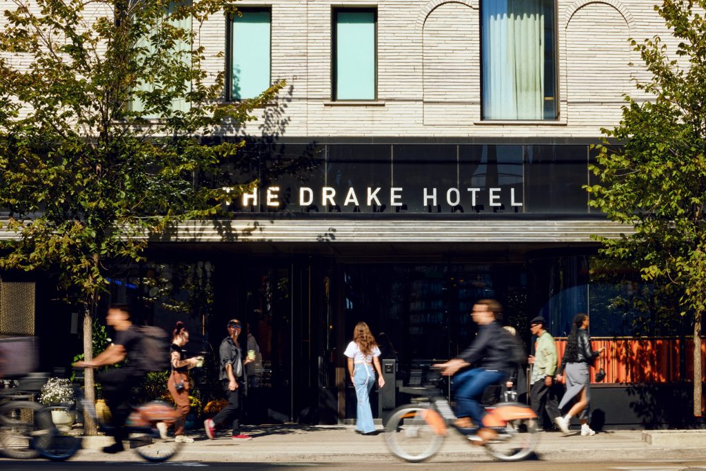 The Drake Hotel | Exterior of the hotel on a sunny day with people walking and biking past and a black awning with "The Drake Hotel" written in white | Homes Almanac