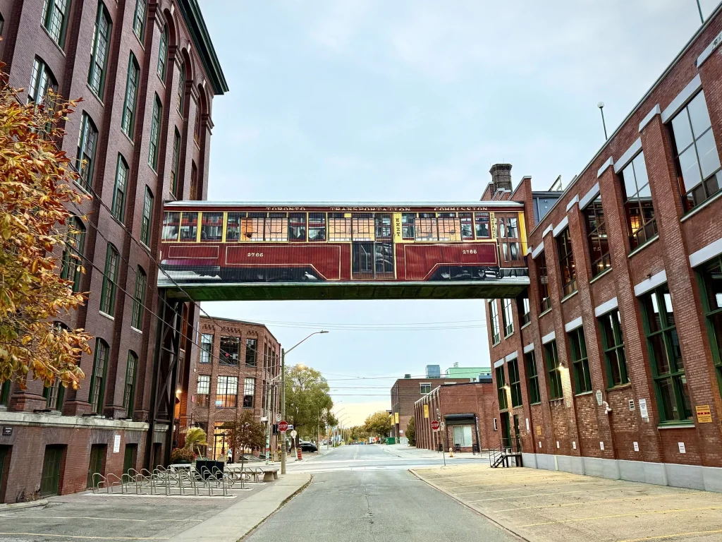 History of Liberty Village | Mural of streetcar on a sky-bridge connecting the Toronto Carpet Factory and the King-Dufferin Corner, of red brick buildings | Homes Almanac
