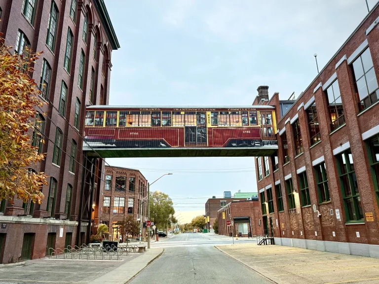 History of Liberty Village | Mural of streetcar on a sky-bridge connecting the Toronto Carpet Factory and the King-Dufferin Corner, of red brick buildings | Homes Almanac