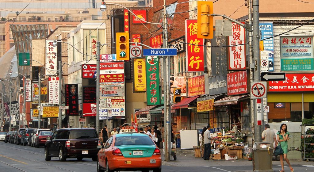 Toronto Chinatown History | A sunny day with brightly coloured signs in Chinese and English off the side of buildings and a taxi driving down the street | Homes Almanac