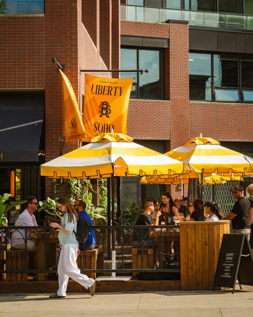 Liberty Soho | Yellow patio umbrellas on a sunny day outside a brick building and people walking and sitting | Homes Almanac