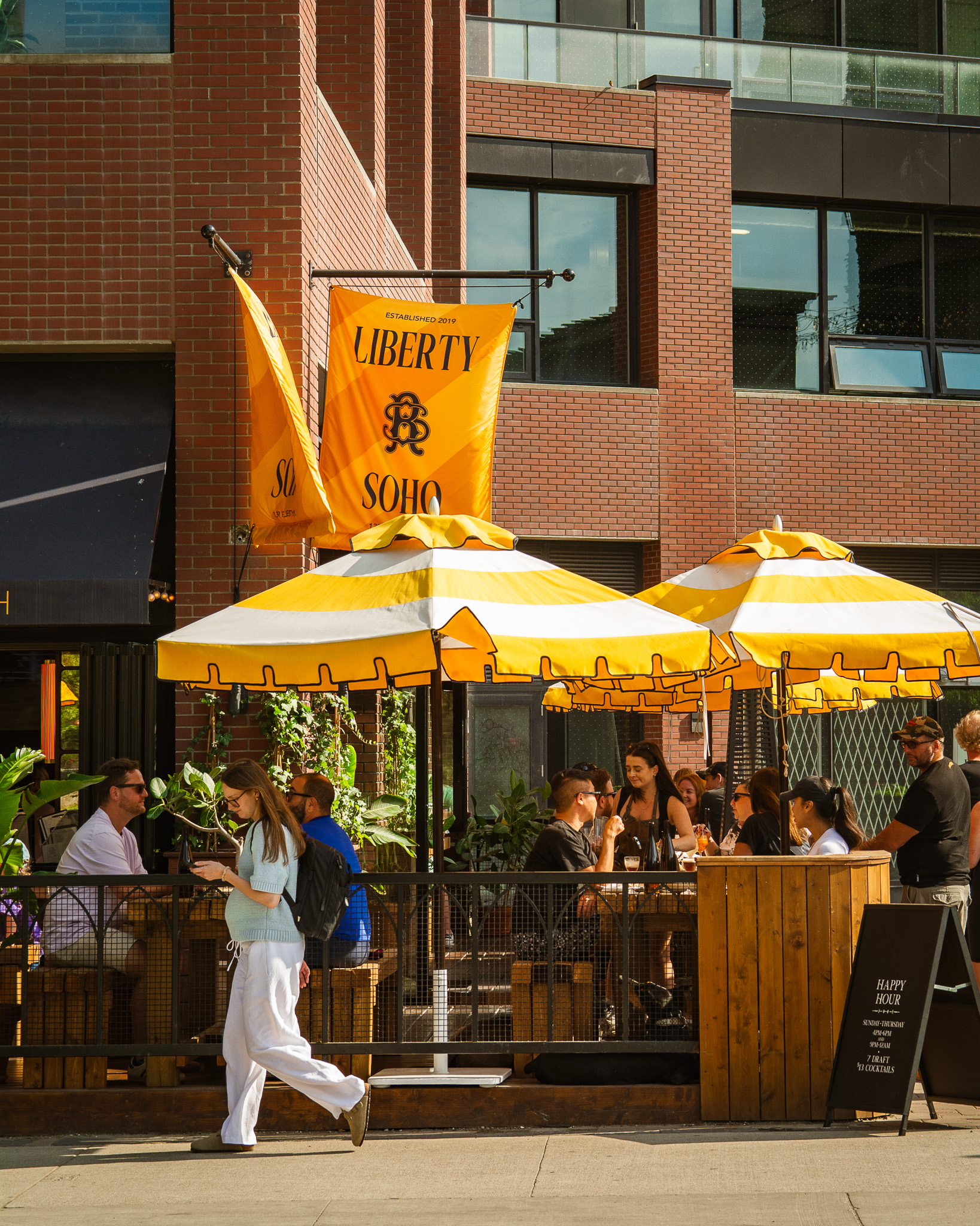Liberty Soho | Yellow patio umbrellas on a sunny day outside a brick building and people walking and sitting | Homes Almanac