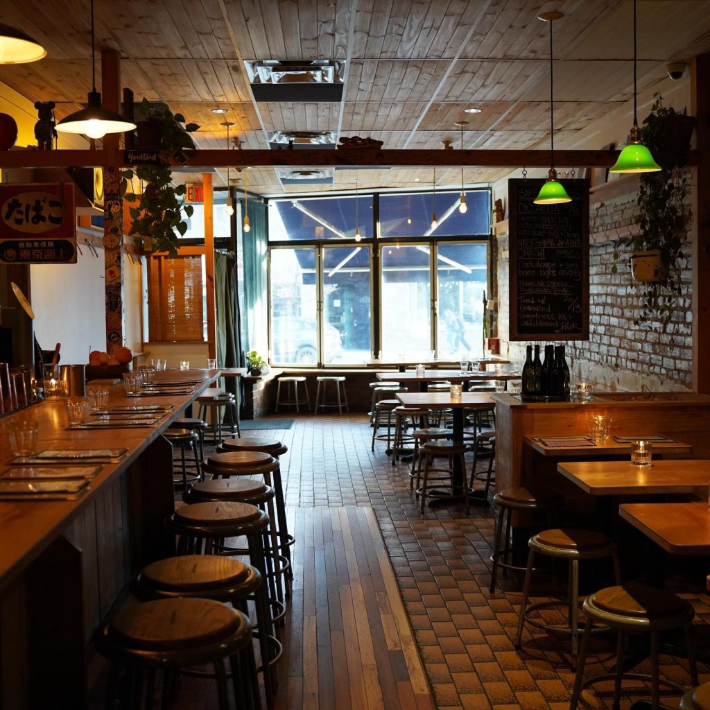 Japanese izakaya toronto | Low lit room with warm wood tables, seating, and bar. Green pot lights hang from the ceiling and brick walls | Homes Almanac