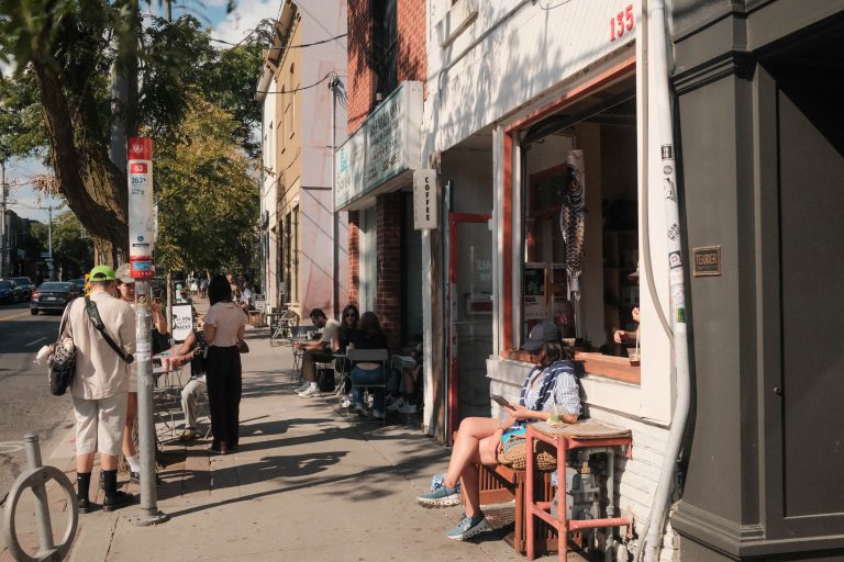 trinity bellwoods 15 minute city | a sunny street with people walking on the sidewalk and sitting outside a coffee shop | homes almanac