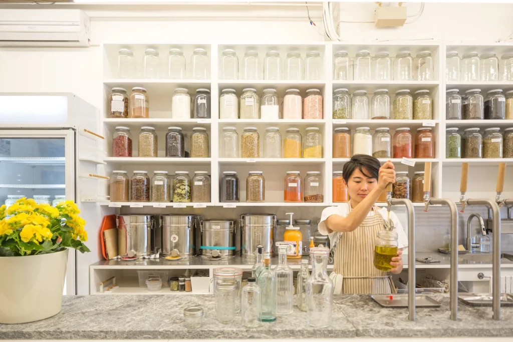 The Soap Dispensary & Kitchen Staples | A woman pouring a liquid from a dispenser with a wall of refillable products on shelves behind her | Homes Almanac